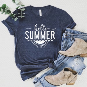 May include: A navy blue t-shirt with the text "hello summer" in white lettering. The text is surrounded by a graphic of a sun and waves. The t-shirt is laid out on a white surface with a pair of blue jeans and brown ankle boots.