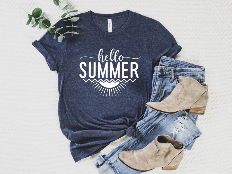 May include: A navy blue t-shirt with the text "hello summer" in white lettering. The text is surrounded by a graphic of a sun and waves. The t-shirt is laid out on a white surface with a pair of blue jeans and brown ankle boots.