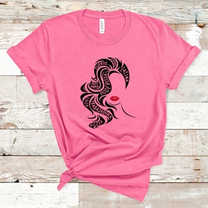 May include: Pink t-shirt with a black graphic design of a woman's face. The design is made up of words in Spanish, including "fuerte", "poderosa", "dama", "trabajadora", "mujer", "latina", and "con."