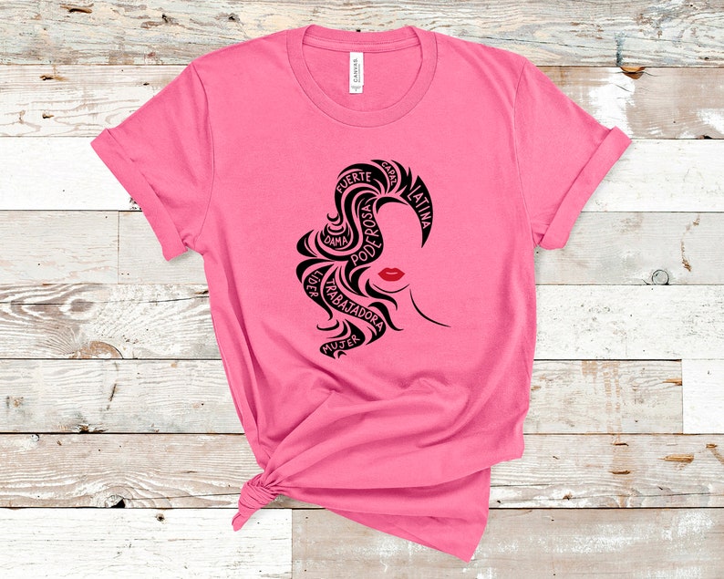 May include: Pink t-shirt with a black graphic design of a woman's face. The design is made up of words in Spanish, including "fuerte", "poderosa", "dama", "trabajadora", "mujer", "latina", and "con."