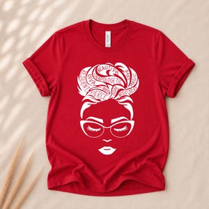 May include: A red short-sleeved t-shirt featuring a white graphic of a face with glasses and a messy bun hairstyle. The hair design incorporates the words beautiful, love, magic, resilient, diva, unapologetic, fierce, queen, dope, boss, and sexy.