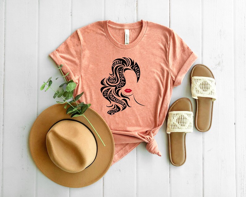 May include: A peach-colored t-shirt with a black silhouette of a woman's head with the words "fuerte", "bella", "poderosa", "trabajadora", "mujer", "latina", "guerrera", "unica", "hermosa", "alma" written around her hair.