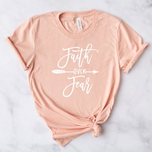 May include: A peach-colored t-shirt with the text "Faith over Fear" in white lettering with a small feather and arrow graphic.