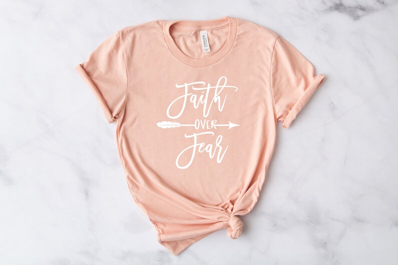May include: A peach-colored t-shirt with the text "Faith over Fear" in white lettering with a small feather and arrow graphic.