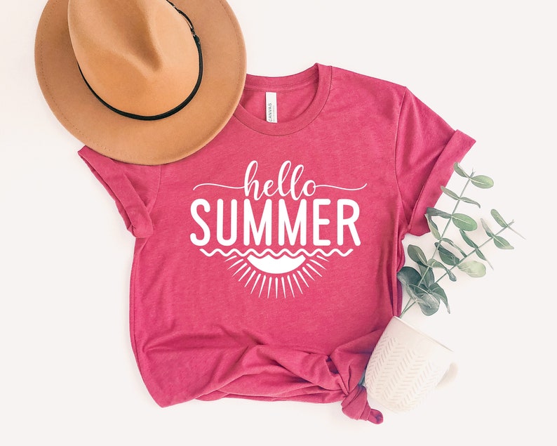 May include: A pink t-shirt with the text "hello summer" in white lettering. The shirt is styled with a brown hat and a sprig of greenery.