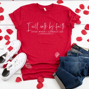 May include: A red t-shirt with the text "I will walk by faith EVEN WHEN I CANNOT SEE 2 CORINTHIANS 5:7" in white script. The shirt is paired with blue jeans and white sneakers. Red rose petals are scattered around the items.