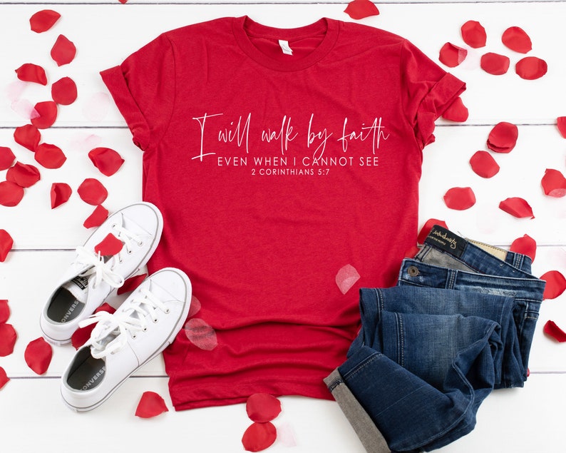 May include: A red t-shirt with the text "I will walk by faith EVEN WHEN I CANNOT SEE 2 CORINTHIANS 5:7" in white script. The shirt is paired with blue jeans and white sneakers. Red rose petals are scattered around the items.