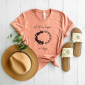 May include: Peach-coloured t-shirt with the text "A lot can happen in 3 days" and a black graphic of a crown of thorns and a wreath. A brown fedora hat and a pair of cream-coloured sandals are also in the image.