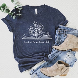May include: A navy blue t-shirt with a white outline of an open book with flowers growing out of it. The text "Custom Name Book Club" is printed below the book.