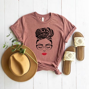 May include: A mauve short-sleeve t-shirt featuring a black graphic of a face with glasses and a bun hairstyle formed by words including beautiful, resilient, and magic, with bright red lips. A tan fedora and two beige woven slide sandals.