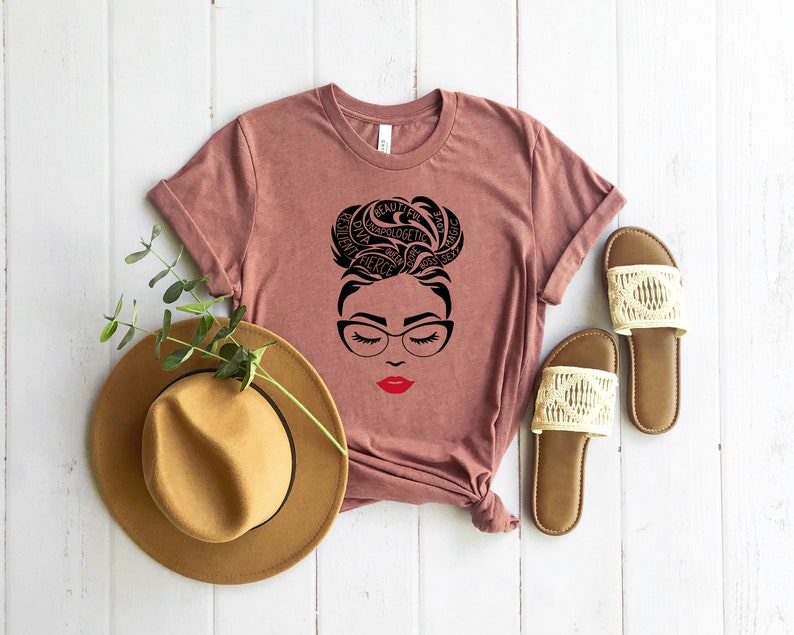 May include: A mauve short-sleeve t-shirt featuring a black graphic of a face with glasses and a bun hairstyle formed by words including beautiful, resilient, and magic, with bright red lips. A tan fedora and two beige woven slide sandals.