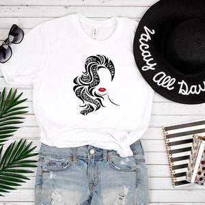 May include: White t-shirt with a black and red graphic of a woman's face with the words "fuerte", "poderosa", "guerrera", "trabajadora", "mujer", "dama", "unica", "latina" written in her hair.