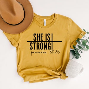May include: A mustard yellow t-shirt with black text that reads "She is Strong" with a line through the word "is" and "Proverbs 31:25" below.