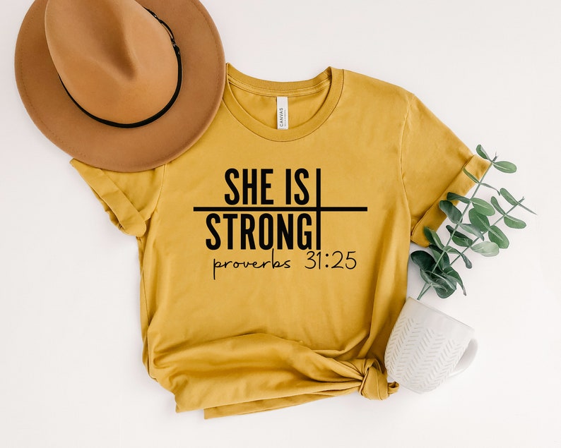 May include: A mustard yellow t-shirt with black text that reads "She is Strong" with a line through the word "is" and "Proverbs 31:25" below.