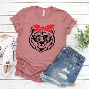 以下が含まれることがあります： ダスティローズのTシャツ。サングラスと赤いバンダナをつけたクマの黒いグラフィック。サングラスの中に「MAMA BEAR」の文字。白いキャンバススニーカーとデニムショーツも画像にあります。