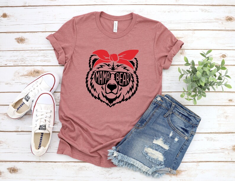 以下が含まれることがあります： ダスティローズのTシャツ。サングラスと赤いバンダナをつけたクマの黒いグラフィック。サングラスの中に「MAMA BEAR」の文字。白いキャンバススニーカーとデニムショーツも画像にあります。