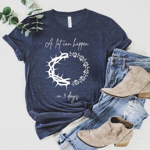 May include: Navy blue t-shirt with white text reading "A lot can happen in 3 days" and a graphic of a crown of thorns and a wreath. The shirt is styled with blue jeans and tan ankle boots.