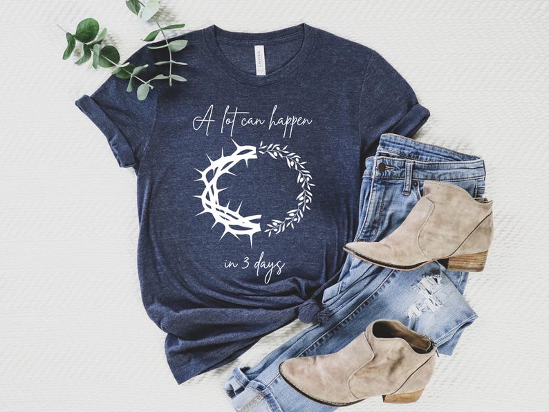 May include: Navy blue t-shirt with white text reading "A lot can happen in 3 days" and a graphic of a crown of thorns and a wreath. The shirt is styled with blue jeans and tan ankle boots.
