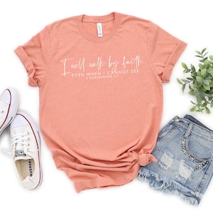 May include: A peach-colored t-shirt with the text "I will walk by faith, even when I cannot see, 2 Corinthians 5:7" in white script. The shirt is paired with denim shorts and white canvas sneakers.