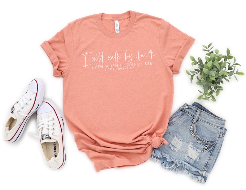 May include: A peach-colored t-shirt with the text "I will walk by faith, even when I cannot see, 2 Corinthians 5:7" in white script. The shirt is paired with denim shorts and white canvas sneakers.