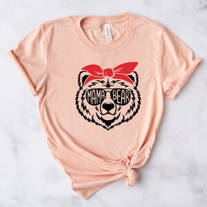 以下が含まれることがあります： サングラスと赤いバンダナをつけたクマの顔のグラフィックが特徴の、明るいピーチ色のTシャツ。「MAMA BEAR」の文字がサングラスの中に表示されています。シャツは柔らかい素材でできており、袖はロールアップされています。