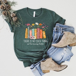 May include: A green t-shirt with a stack of colorful books and flowers printed on the front. The text "THERE IS NO SUCH THING as too many books" is printed below the books. The t-shirt is laid out on a white surface with a pair of blue jeans and brown ankle boots.
