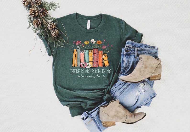 May include: A green t-shirt with a stack of colorful books and flowers printed on the front. The text "THERE IS NO SUCH THING as too many books" is printed below the books. The t-shirt is laid out on a white surface with a pair of blue jeans and brown ankle boots.