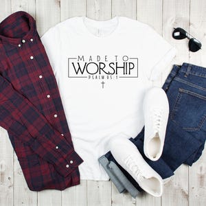 Puede incluir: Camiseta blanca con el texto "MADE TO WORSHIP - PSALM 95:1" en letras negras. La camiseta est&aacute; colocada sobre una superficie de madera con una camisa de cuadros, pantalones vaqueros y zapatillas blancas.