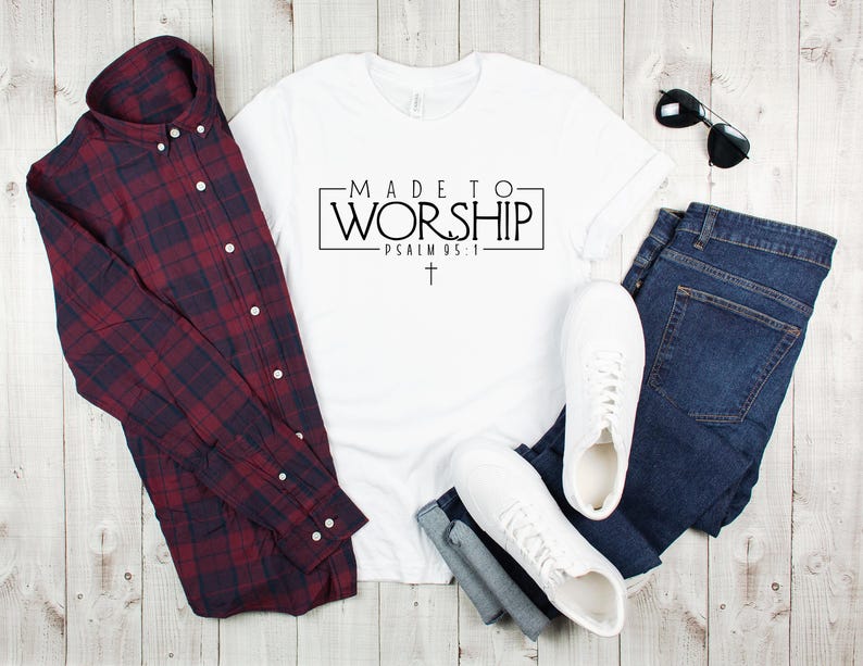 Puede incluir: Camiseta blanca con el texto "MADE TO WORSHIP - PSALM 95:1" en letras negras. La camiseta est&aacute; colocada sobre una superficie de madera con una camisa de cuadros, pantalones vaqueros y zapatillas blancas.