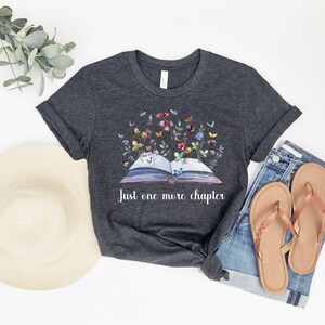 Op de afbeelding: Een donkergrijs t-shirt met een afbeelding van een open boek met bloemen en vlinders die eruit groeien. De tekst "Just one more chapter" staat onder het boek.