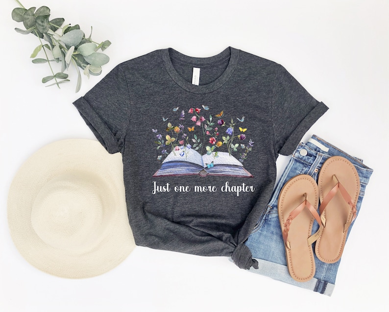 Op de afbeelding: Een donkergrijs t-shirt met een afbeelding van een open boek met bloemen en vlinders die eruit groeien. De tekst "Just one more chapter" staat onder het boek.