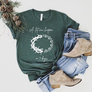 May include: Dark green t-shirt with the text "A lot can happen in 3 days" and a white graphic of a crown of thorns and a wreath. The shirt is paired with blue jeans and tan ankle boots.