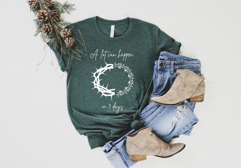 May include: Dark green t-shirt with the text "A lot can happen in 3 days" and a white graphic of a crown of thorns and a wreath. The shirt is paired with blue jeans and tan ankle boots.