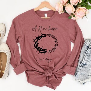 May include: A long-sleeved, mauve-coloured shirt with the text "A lot can happen in 3 days" and a black graphic of a crown of thorns and a wreath. The shirt is displayed with cream-coloured flats and denim shorts.