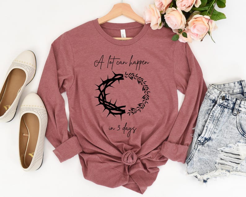May include: A long-sleeved, mauve-coloured shirt with the text "A lot can happen in 3 days" and a black graphic of a crown of thorns and a wreath. The shirt is displayed with cream-coloured flats and denim shorts.