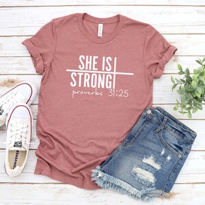 May include: A dusty rose t-shirt with the text "SHE IS STRONG" with a line through the "IS" and the text "proverbs 31:25" below.