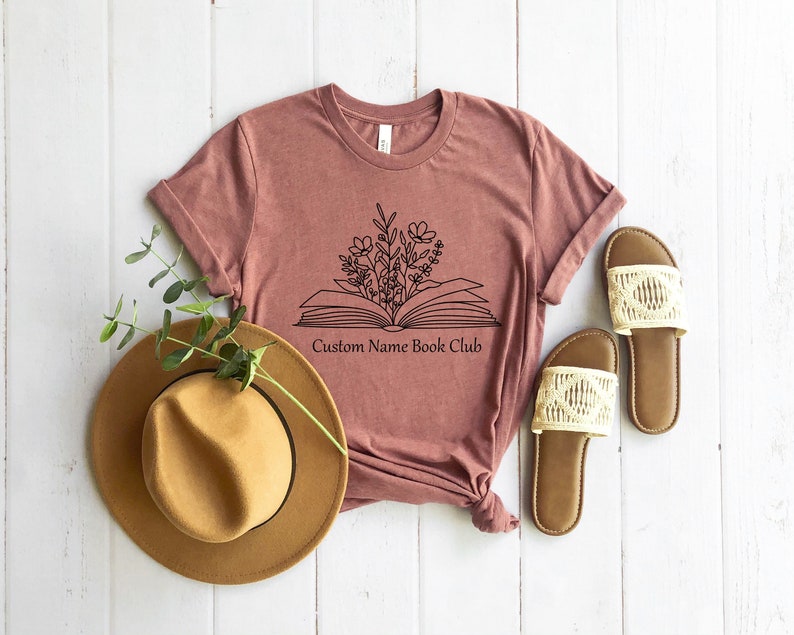 May include: A mauve t-shirt with a black outline of an open book with flowers growing out of it. The text "Custom Name Book Club" is printed below the book.