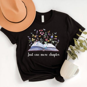 Op de afbeelding: Zwart T-shirt met een afbeelding van een open boek met bloemen en vlinders. De tekst "Just one more chapter" staat onder het boek.