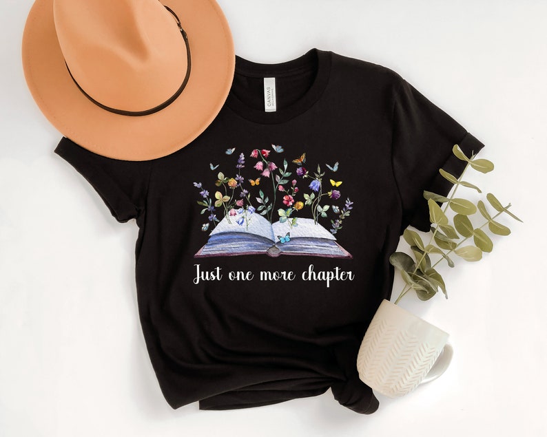 Op de afbeelding: Zwart T-shirt met een afbeelding van een open boek met bloemen en vlinders. De tekst "Just one more chapter" staat onder het boek.