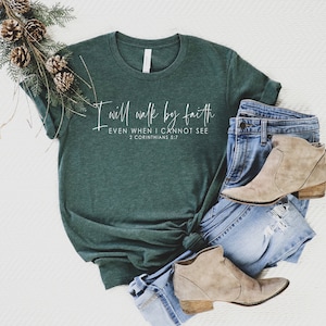 May include: A dark green t-shirt with the text "I will walk by faith, even when I cannot see, 2 Corinthians 5:7" in white script. The shirt is paired with blue jeans and tan ankle boots. A decorative pine branch with pinecones is placed on the side.