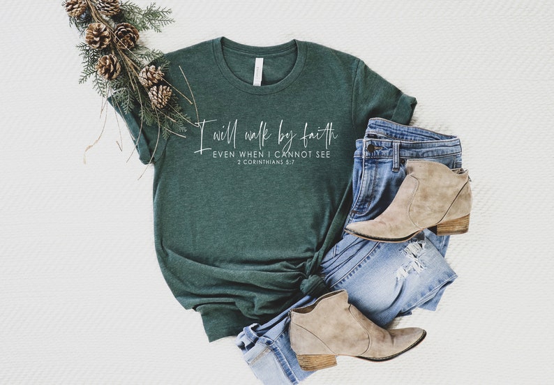May include: A dark green t-shirt with the text "I will walk by faith, even when I cannot see, 2 Corinthians 5:7" in white script. The shirt is paired with blue jeans and tan ankle boots. A decorative pine branch with pinecones is placed on the side.