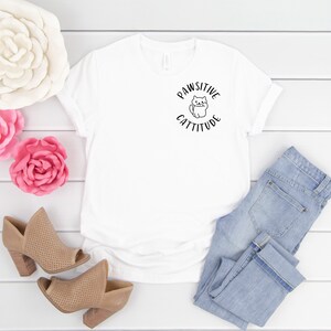 Puede incluir: Camiseta blanca con un gr&aacute;fico negro de un gato y el texto "Pawsitive Cattitude". La camiseta est&aacute; colocada sobre una superficie de madera blanca con un par de zapatos de tac&oacute;n marr&oacute;n, dos flores rosas y un par de pantalones vaqueros azules claros.