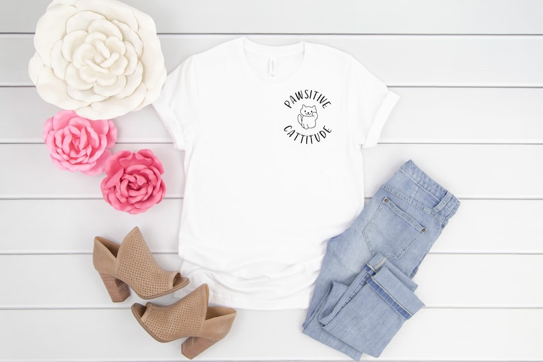 Puede incluir: Camiseta blanca con un gr&aacute;fico negro de un gato y el texto "Pawsitive Cattitude". La camiseta est&aacute; colocada sobre una superficie de madera blanca con un par de zapatos de tac&oacute;n marr&oacute;n, dos flores rosas y un par de pantalones vaqueros azules claros.