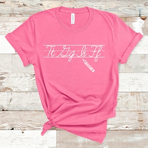 May include: Pink t-shirt with a white graphic of cursive letters "Ft Zg Li Ff" on lined paper with a pencil above the letters.