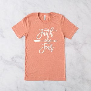 May include: A peach-colored t-shirt with the text "Faith over Fear" in white lettering with a small arrow and feather design.