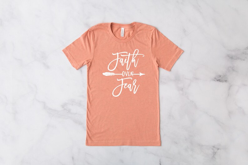 May include: A peach-colored t-shirt with the text "Faith over Fear" in white lettering with a small arrow and feather design.