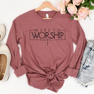 Puede incluir: Una camiseta de manga larga de color granate con el texto "MADE TO WORSHIP - PSALM 95:1" impreso en la parte delantera. Una peque&ntilde;a cruz se encuentra debajo del texto.