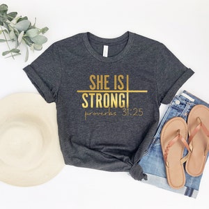 May include: A dark gray t-shirt with gold lettering that reads "She is Strong" with a horizontal line through the word "Strong" and the text "Proverbs 31:25" below.