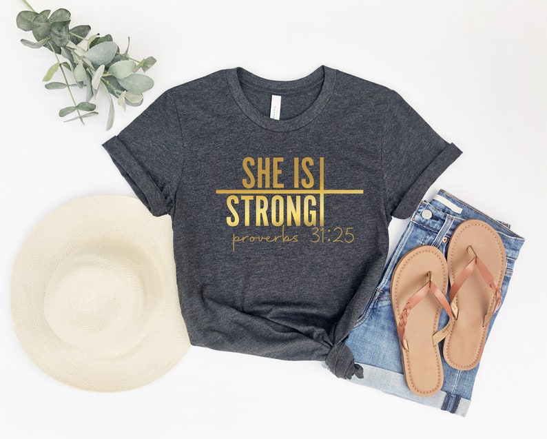 May include: A dark gray t-shirt with gold lettering that reads "She is Strong" with a horizontal line through the word "Strong" and the text "Proverbs 31:25" below.