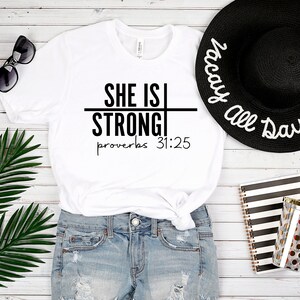 May include: White t-shirt with black text that reads "She is Strong" with a line through the word "Strong" and the text "Proverbs 31:25" below. The t-shirt is worn with denim shorts and a black sun hat with white text that reads "Vacay All Day".